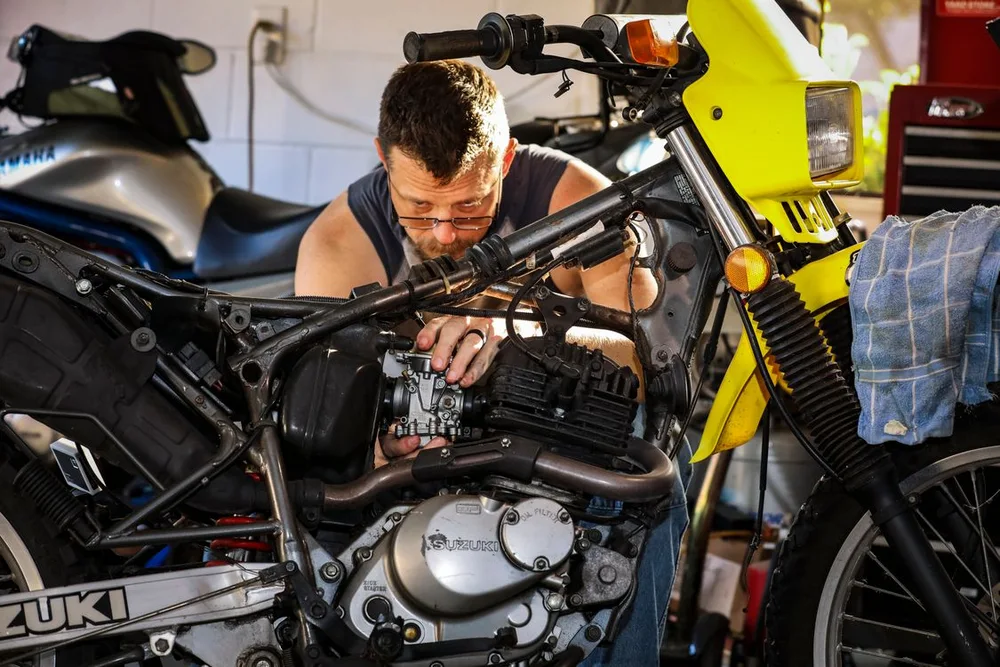 A mechanic works on the carburetor of a motorcycle with its tank removed. Illustrative photo.